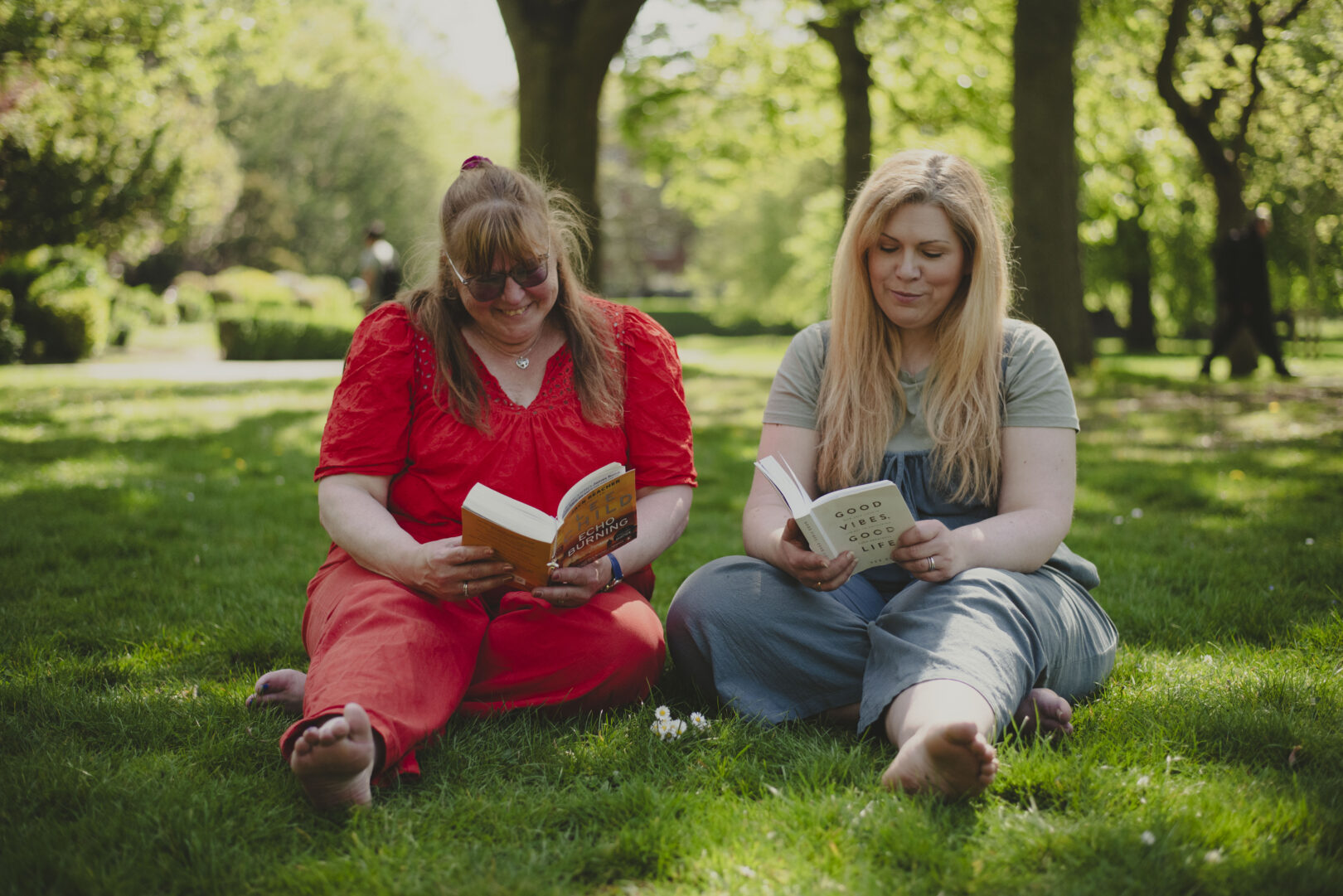 Two people sitting on the grass in a park reading a book. 