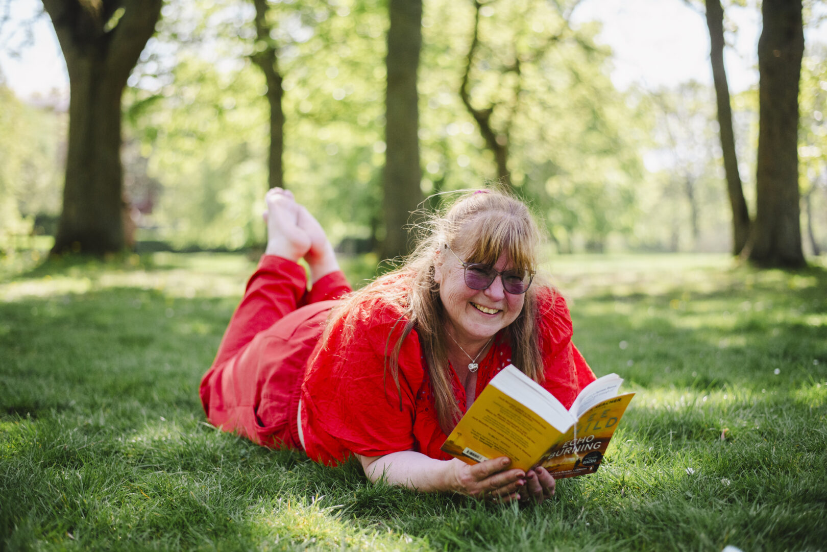 A woman in a red outfit lies on the grass in a park, smiling and reading a book on a sunny day. Image provided courtesy of Leeds Beckett University - leedsbeckett.ac.uk
