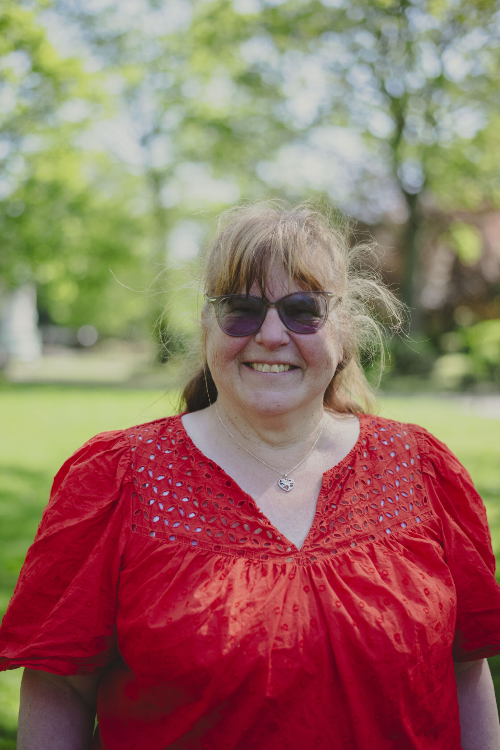 A woman wearing sunglasses and a red blouse is smiling outdoors, with green trees and blurred buildings in the background. Image provided courtesy of Leeds Beckett University - leedsbeckett.ac.uk