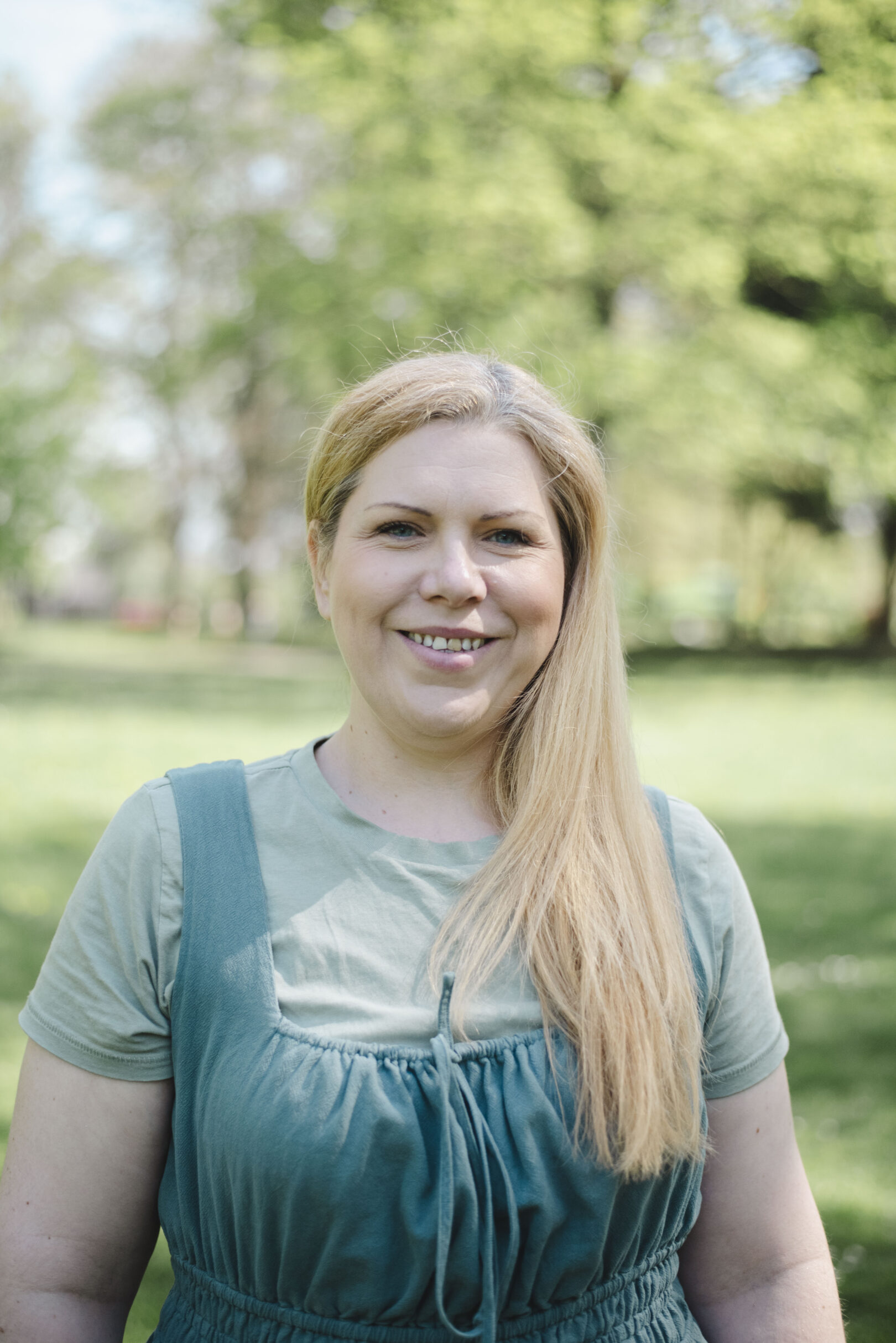 A woman with long blonde hair, wearing a green top and dress, stands outdoors in a park with green trees in the background. Image provided courtesy of Leeds Beckett University - leedsbeckett.ac.uk