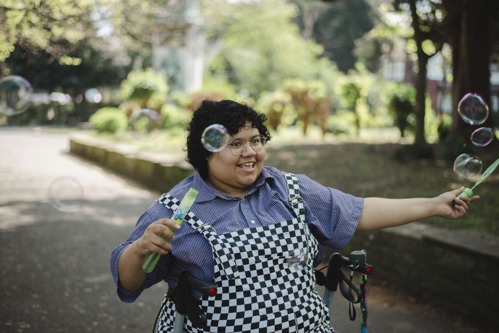 A person wearing a checkered dress and glasses, smiles while blowing bubbles in an outdoor park setting. Image provided courtesy of Leeds Beckett University - leedsbeckett.ac.uk