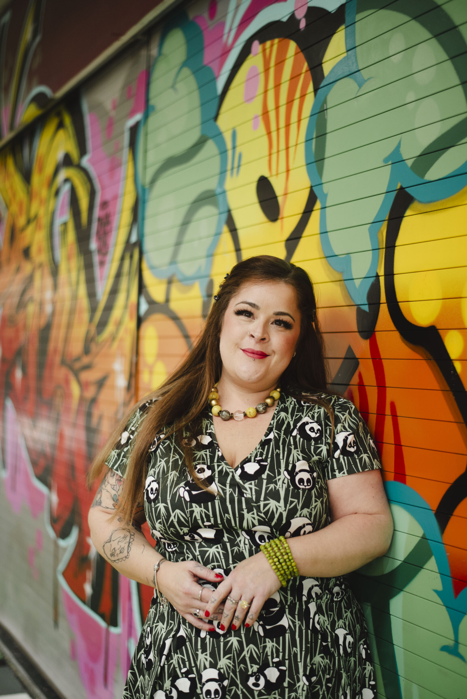 A woman with long hair stands in front of a colorful graffiti-covered wall, wearing a patterned dress and jewelry, with her hands clasped. Image provided courtesy of Leeds Beckett University - leedsbeckett.ac.uk