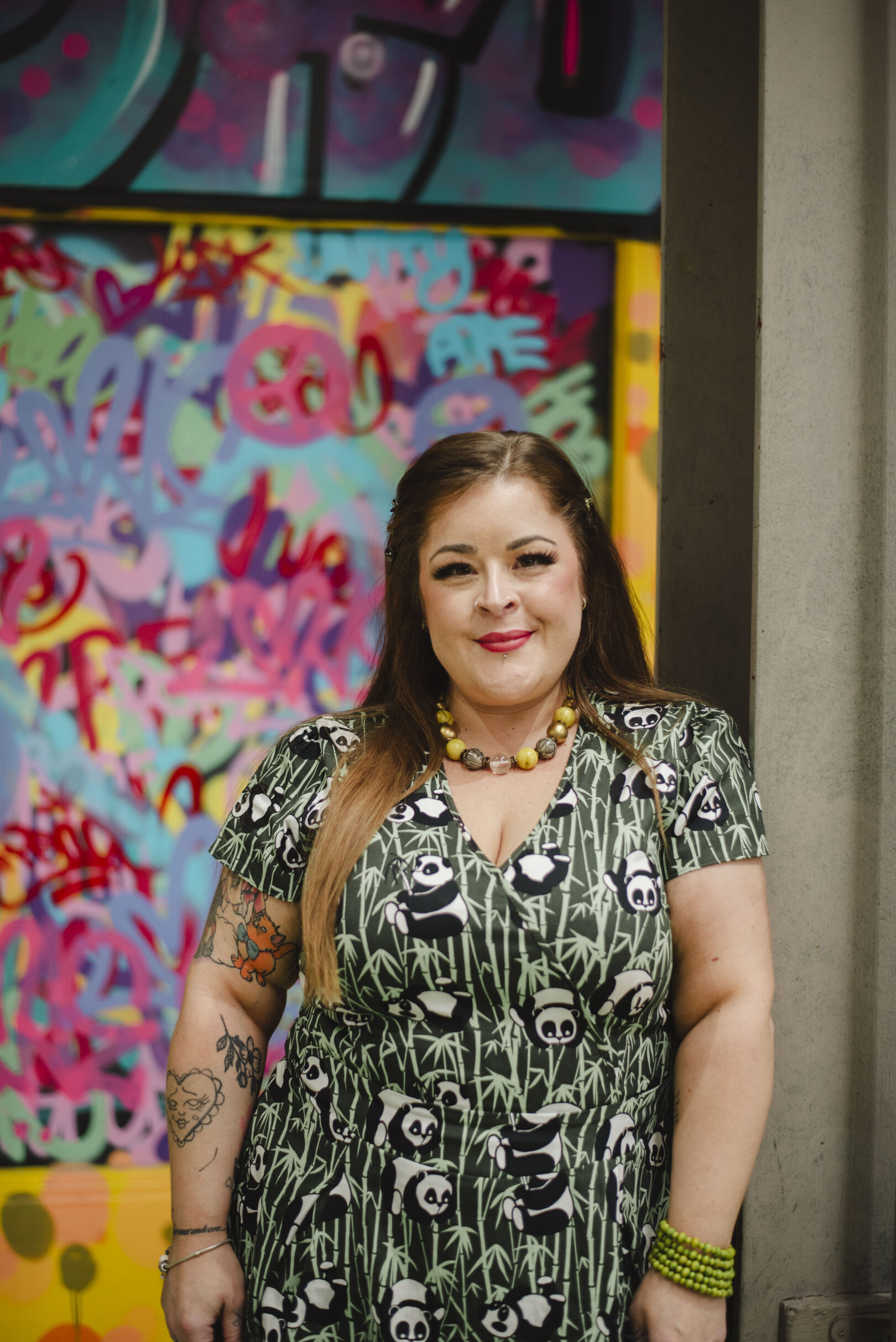 A woman with long hair in a panda print dress stands in front of a colourful graffiti wall, smiling at the camera. Image provided courtesy of Leeds Beckett University - leedsbeckett.ac.uk