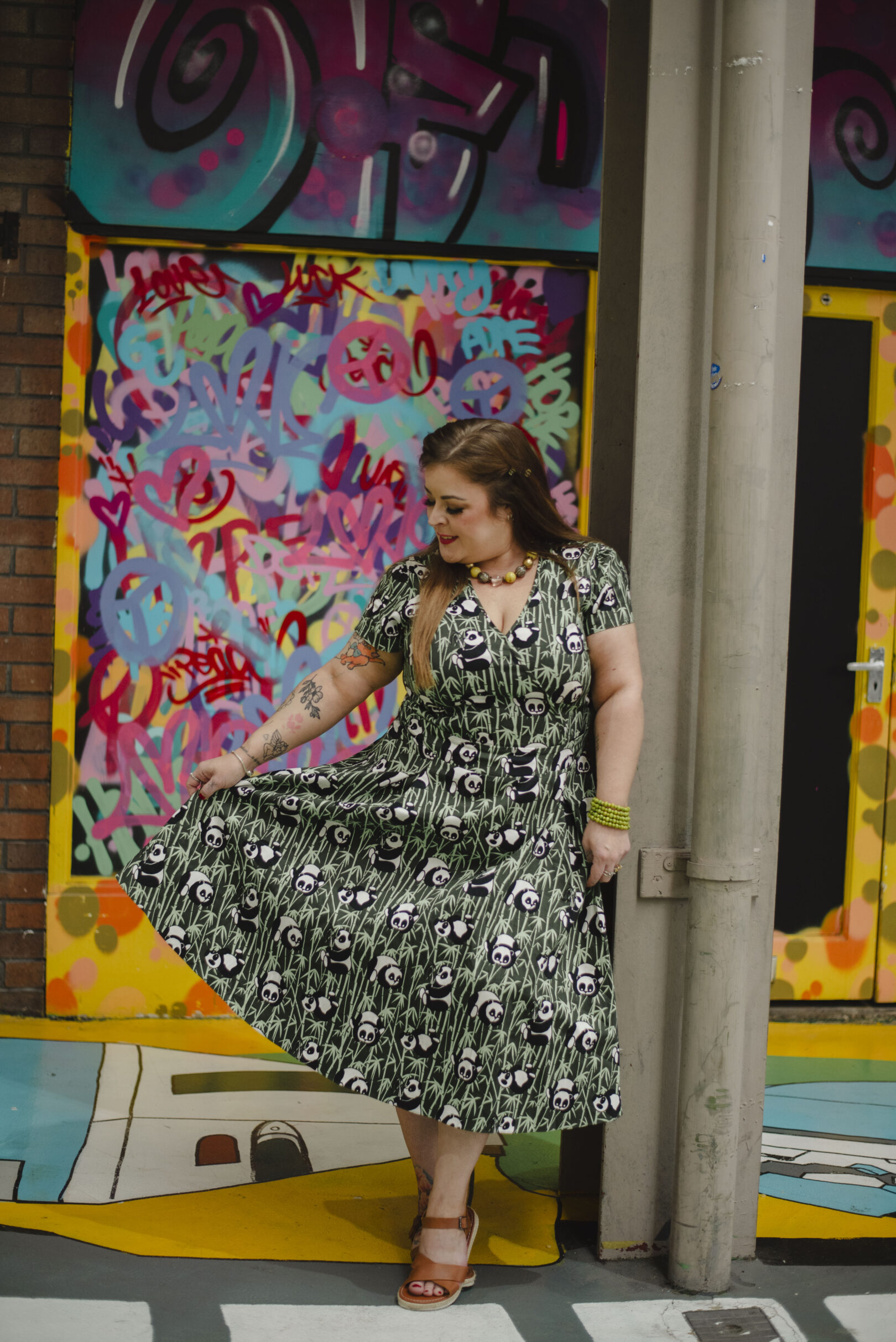 A woman in a patterned green dress stands next to a pole, holding her skirt, with colourful graffiti art in the background. Image provided courtesy of Leeds Beckett University - leedsbeckett.ac.uk